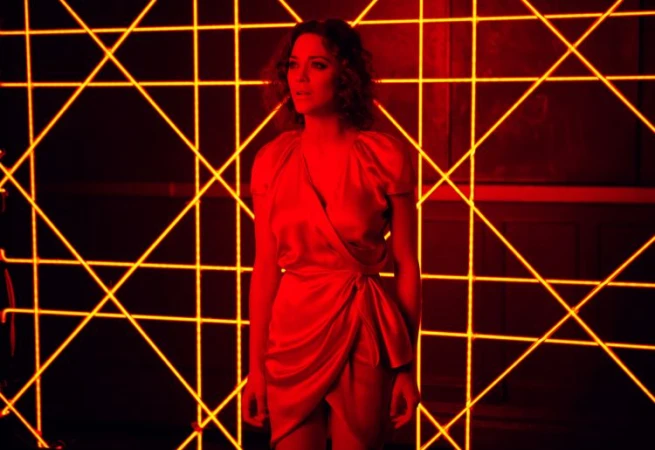 Woman in a red dress stands in front of a wall illuminated with a geometric pattern of yellow neon lights in a studio setting.