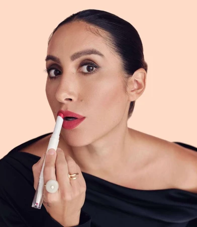 Close-up portrait of a woman applying pink lipstick, wearing a black off-the-shoulder top against a peach background.