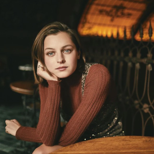 Indoor portrait of an adult woman with blue eyes wearing a brown turtleneck sweater and a sleeveless sequined top sitting at a table.