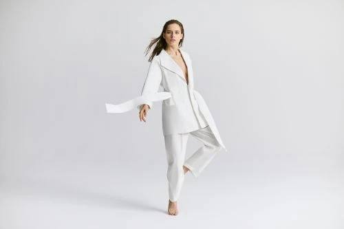 Woman in white suit posing in a studio with one leg up and fabric flowing with wind. Fashion photography for beauty and apparel.