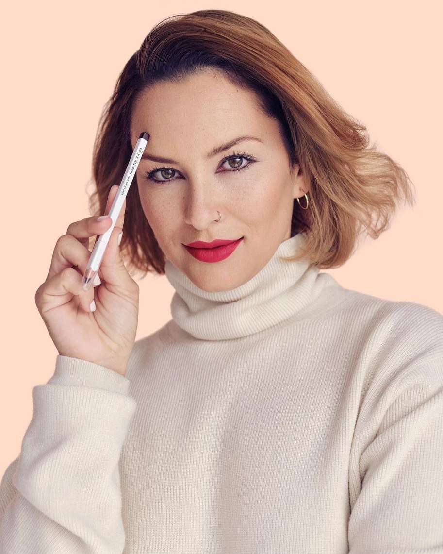 Attractive adult woman wearing a cream-colored turtleneck sweater is holding an eyebrow pencil to her eyebrow in a close-up portrait.
