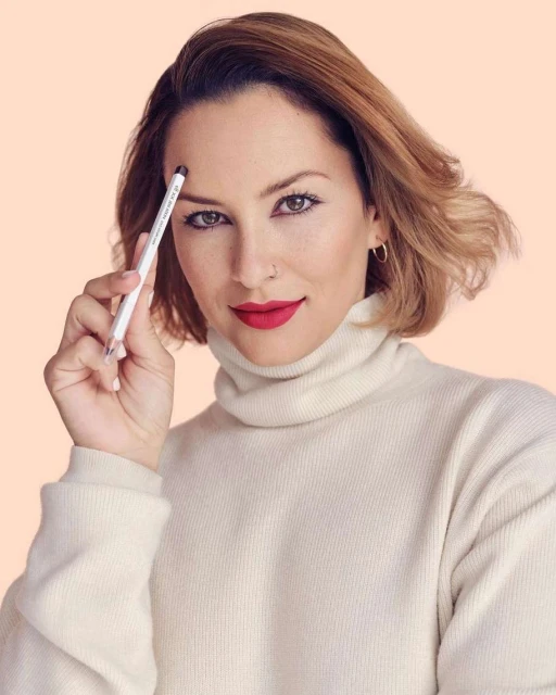 Attractive adult woman wearing a cream-colored turtleneck sweater is holding an eyebrow pencil to her eyebrow in a close-up portrait.