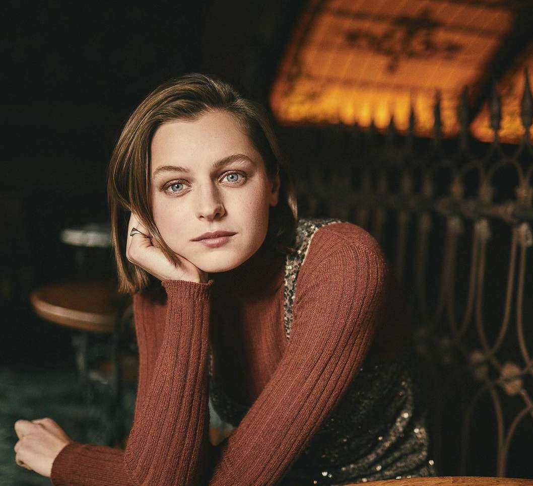 Close-up portrait of a young adult female with bob haircut, wearing a rust-colored turtleneck and a sleeveless sequin top indoors.