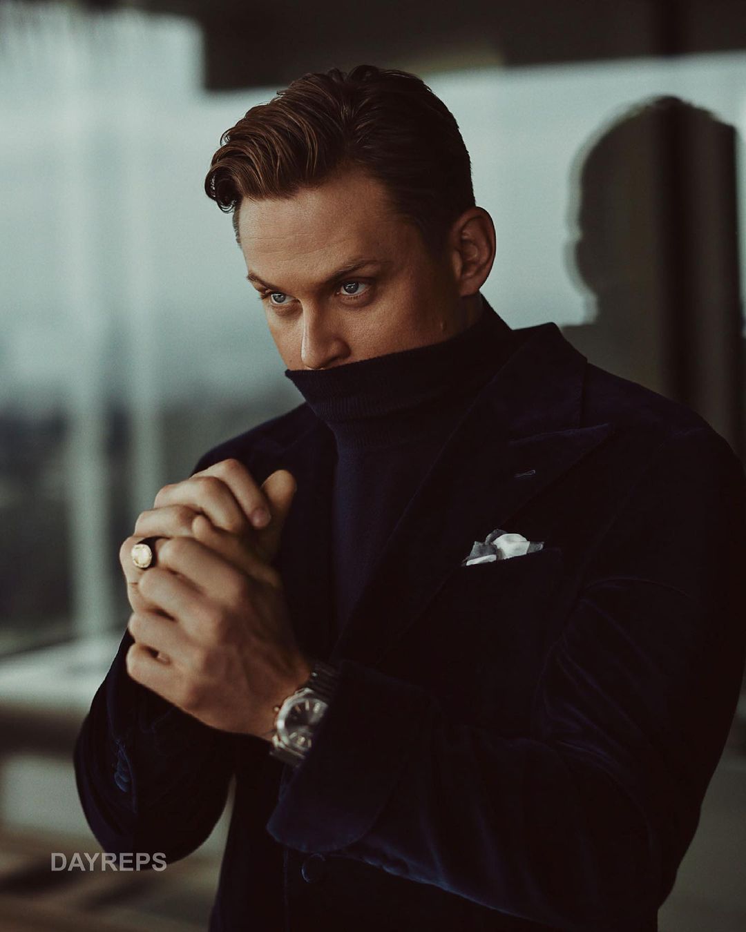 Portrait of a man wearing a dark blue coat, with his hands folded near his face, showcasing his ring and watch in an indoor setting.