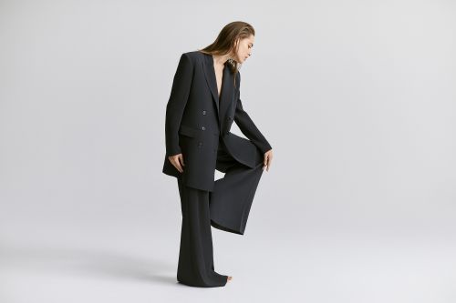 A woman in a black pantsuit strikes a pose on a grey backdrop, one leg bent with her hand resting on the raised knee.