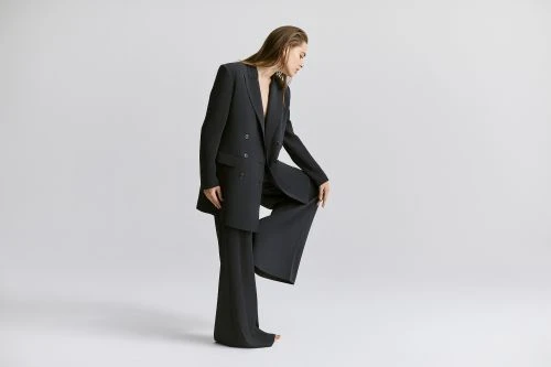 A woman in a black pantsuit strikes a pose on a grey backdrop, one leg bent with her hand resting on the raised knee.