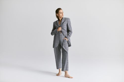 Young woman poses in gray plaid suit, hand in pocket, looking left in bright studio lighting against a clean white backdrop.