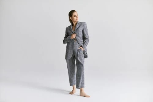 Young woman poses in gray plaid suit, hand in pocket, looking left in bright studio lighting against a clean white backdrop.