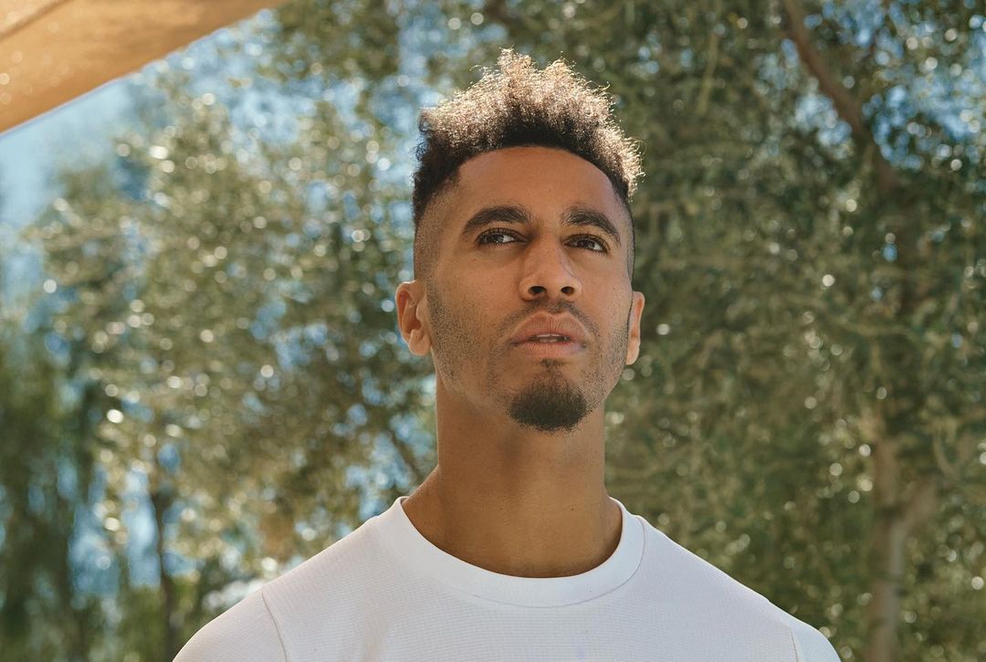 Outdoor headshot of a young adult man with short curly hair and a white t-shirt, looking upward against a bokeh tree background in natural light.