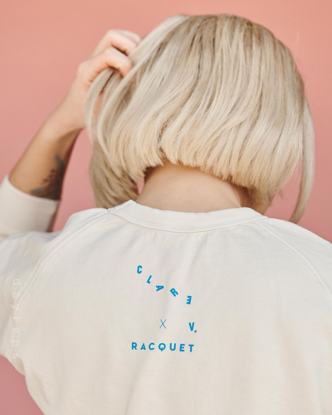 Back view of a woman with a blonde bob haircut wearing a white sweatshirt with the text CLARE V. RACQUET in blue on a peach background.