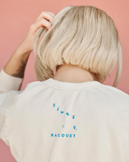 Back view of a woman with a blonde bob haircut wearing a white sweatshirt with the text CLARE V. RACQUET in blue on a peach background.
