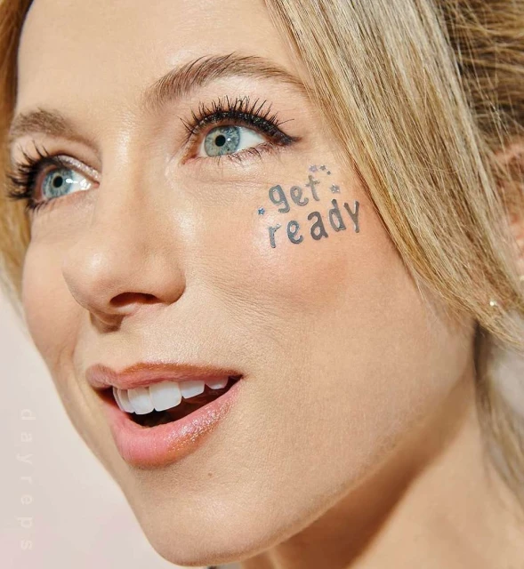 Close-up of a blond woman's face with 'get ready' written on her cheek and glitter accents, creating a beauty or fashion shot.