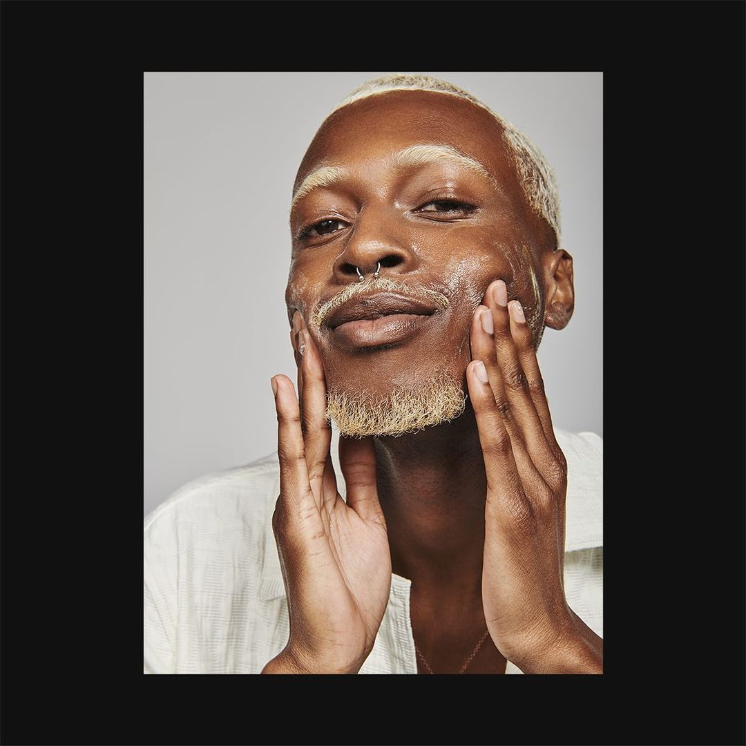 Close-up studio portrait of a Black adult male model with fair hair and beard, applying skincare product to his face with both hands.