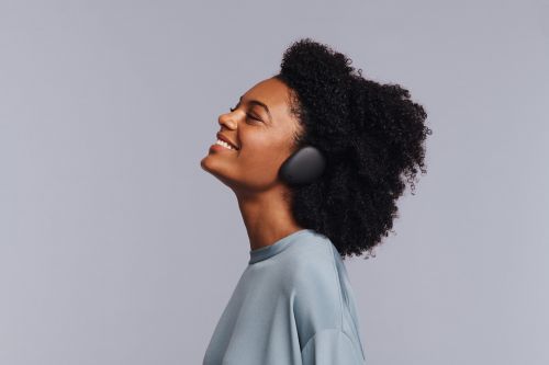Profile view of a smiling adult woman with dark curly hair wearing wireless headphones and a blue t-shirt against a grey background.