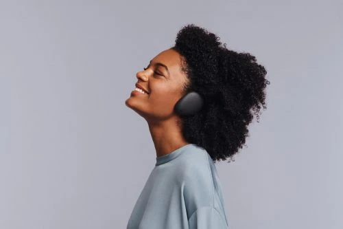 Profile view of a smiling adult woman with dark curly hair wearing wireless headphones and a blue t-shirt against a grey background.
