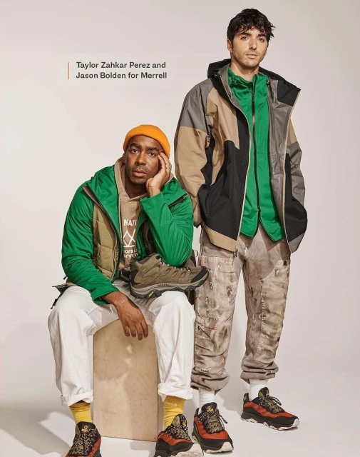 Two men model Merrell apparel and hiking shoes in a studio shot. One man sits on a box wearing a green jacket and an orange beanie.