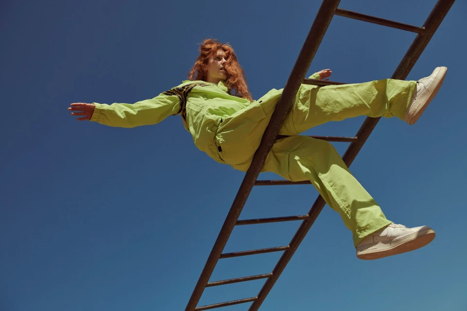 A red-haired adult woman in a lime-green tracksuit balances on a rusted metal ladder against a clear blue sky outdoors in daylight.