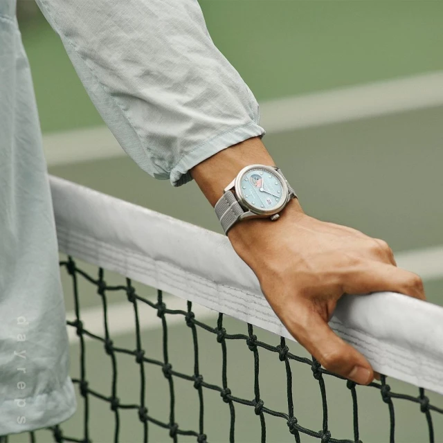 Close-up on a man's wrist wearing a light blue wristwatch with a grey band while holding a tennis net on a green tennis court.