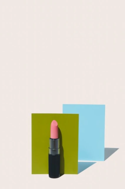 A pink lipstick stands against green and blue squares on a cream background, creating a visually striking beauty and cosmetics still life.