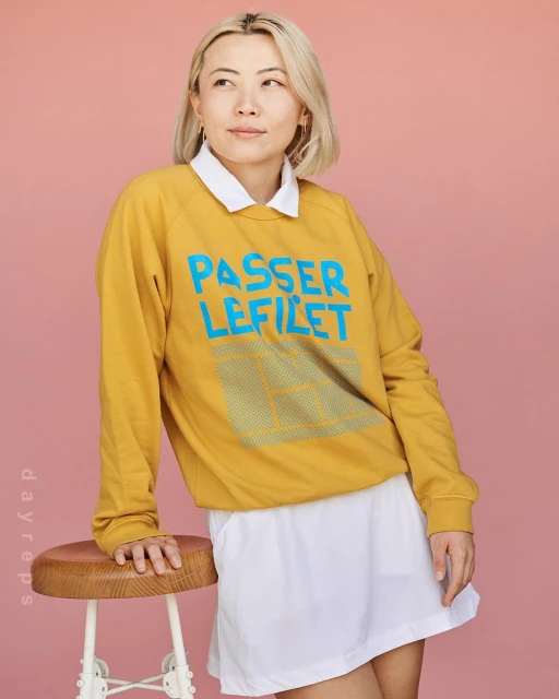 Portrait of a young adult woman with blonde hair and a yellow 'Passer Lefilet' sweatshirt standing next to a brown wooden stool.