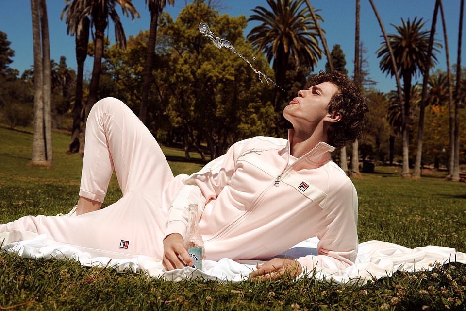 Athletic man reclines on white cloth in sunny park, squirting mineral water in air; wearing pink Fila tracksuit, palm trees.