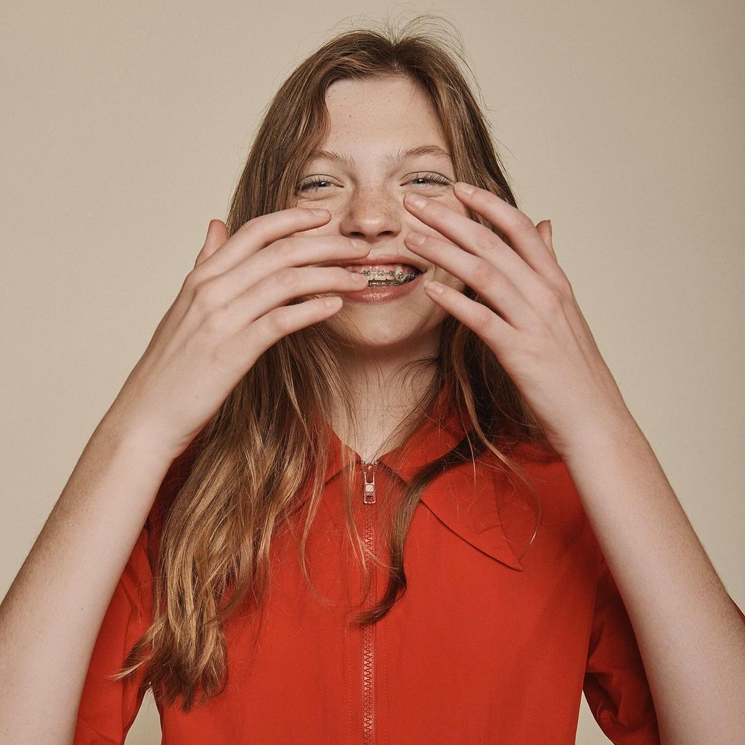 A young woman with long blonde hair smiles, covering her face with her hands, in a close-up studio portrait against a beige background.