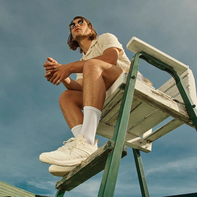 Low angle view of a man with sunglasses sitting on a lifeguard chair, looking off into the distance with a cloudy blue sky background.