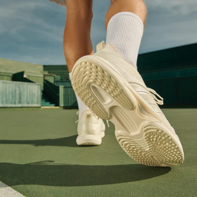 Close-up of a person wearing a white sneaker on a green tennis court with a blurred background of green walls and a blue sky above.