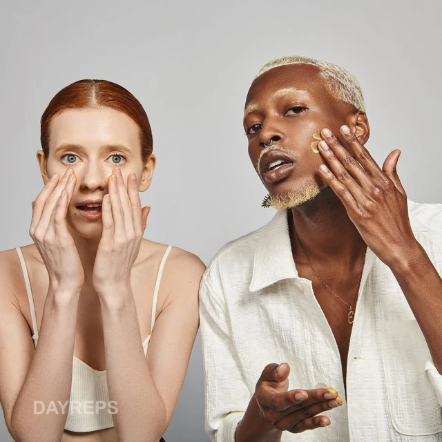 Two diverse young adults apply skincare. A blonde male in a shirt and a red-haired female wear minimal makeup against a grey backdrop.