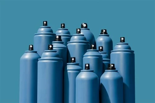 Many blue spray paint cans are grouped together on a blue background in a studio, creating a visually striking monochrome arrangement.