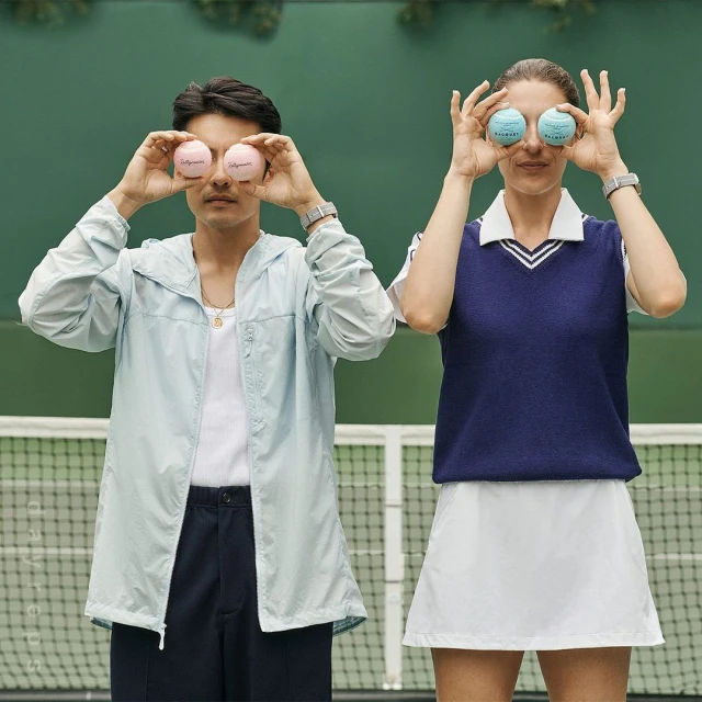 Two people posing with tennis balls in front of their eyes in front of a green wall, wearing tennis outfits, on an outdoor tennis court.