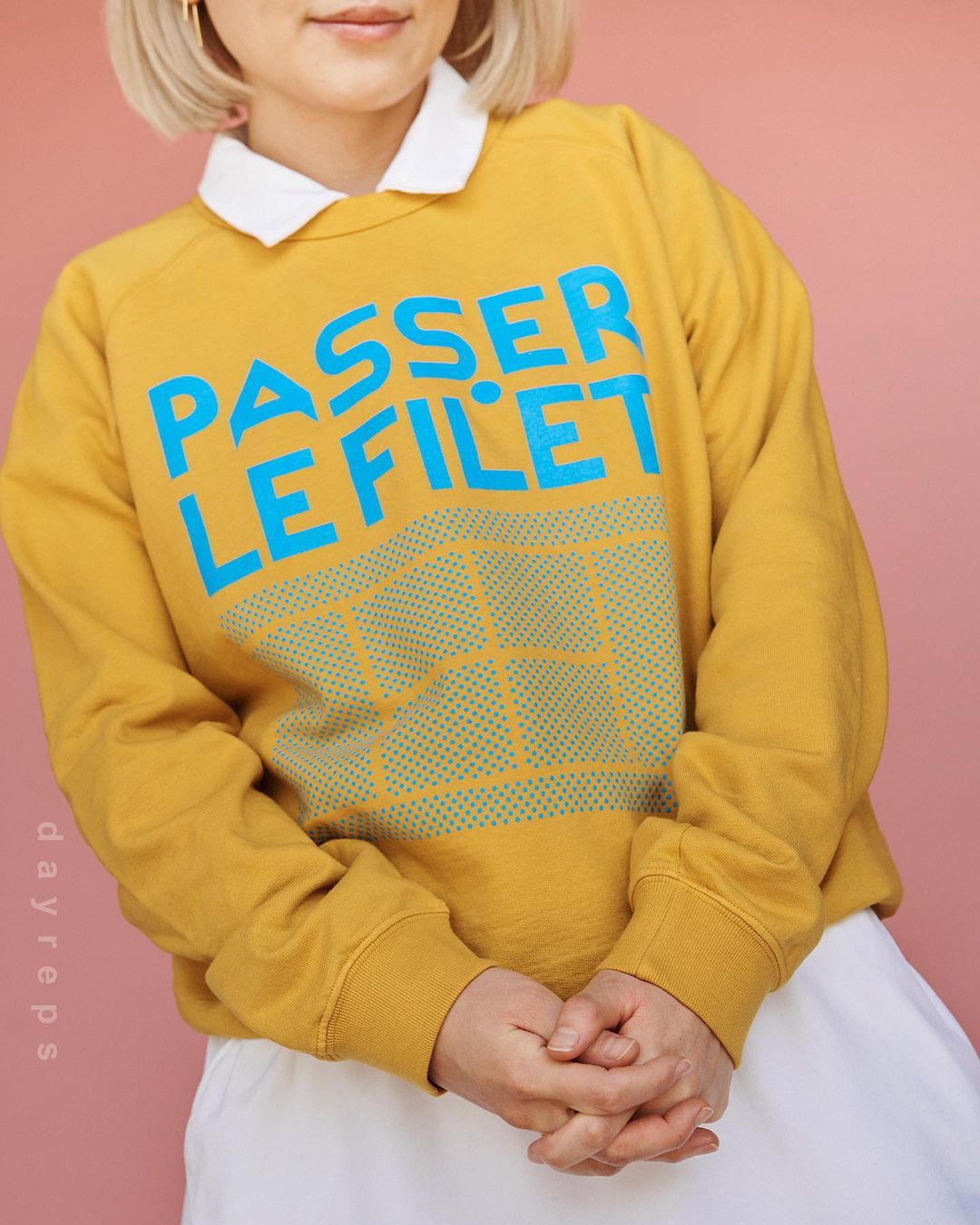 Woman poses indoors wearing mustard-yellow sweatshirt with the words "PASSER LE FILET" in turquoise lettering.