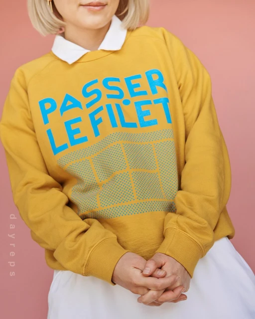 Woman poses indoors wearing mustard-yellow sweatshirt with the words "PASSER LE FILET" in turquoise lettering.