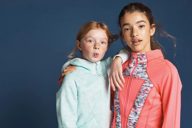 Two young girls posing together, one in a mint green hoodie and one in a bright coral zip jacket against a dark blue background.
