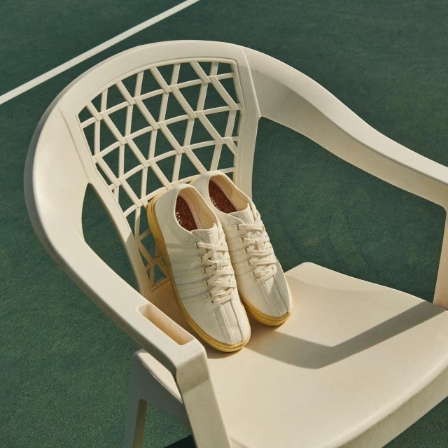 A pair of cream Keds sneakers sits on a white plastic chair on a green tennis court, bathed in bright sunlight.