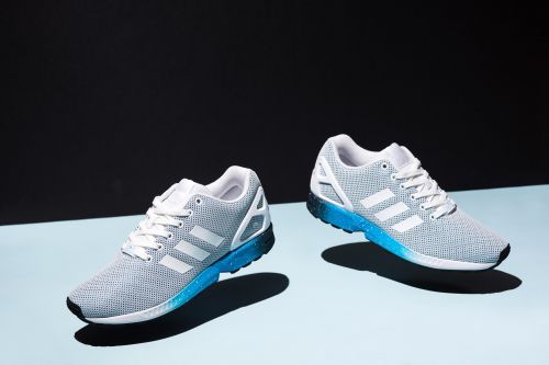 Two gray Adidas running shoes with white stripes and blue soles are featured against a split blue and black background in a studio setting.