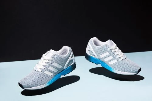 Two gray Adidas running shoes with white stripes and blue soles are featured against a split blue and black background in a studio setting.