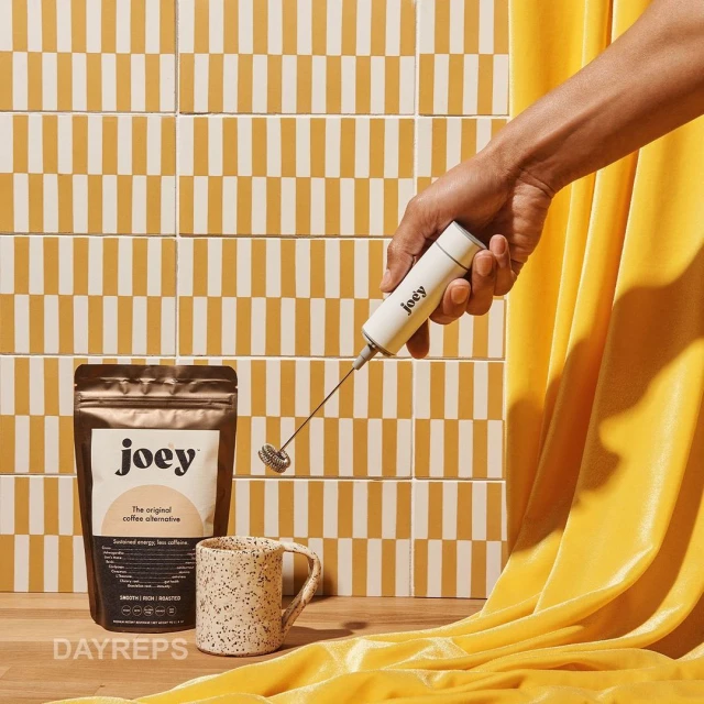 Hand holding Joey coffee alternative mixer, a bag of Joey coffee alternative, and a mug on a yellow and white tiled surface.