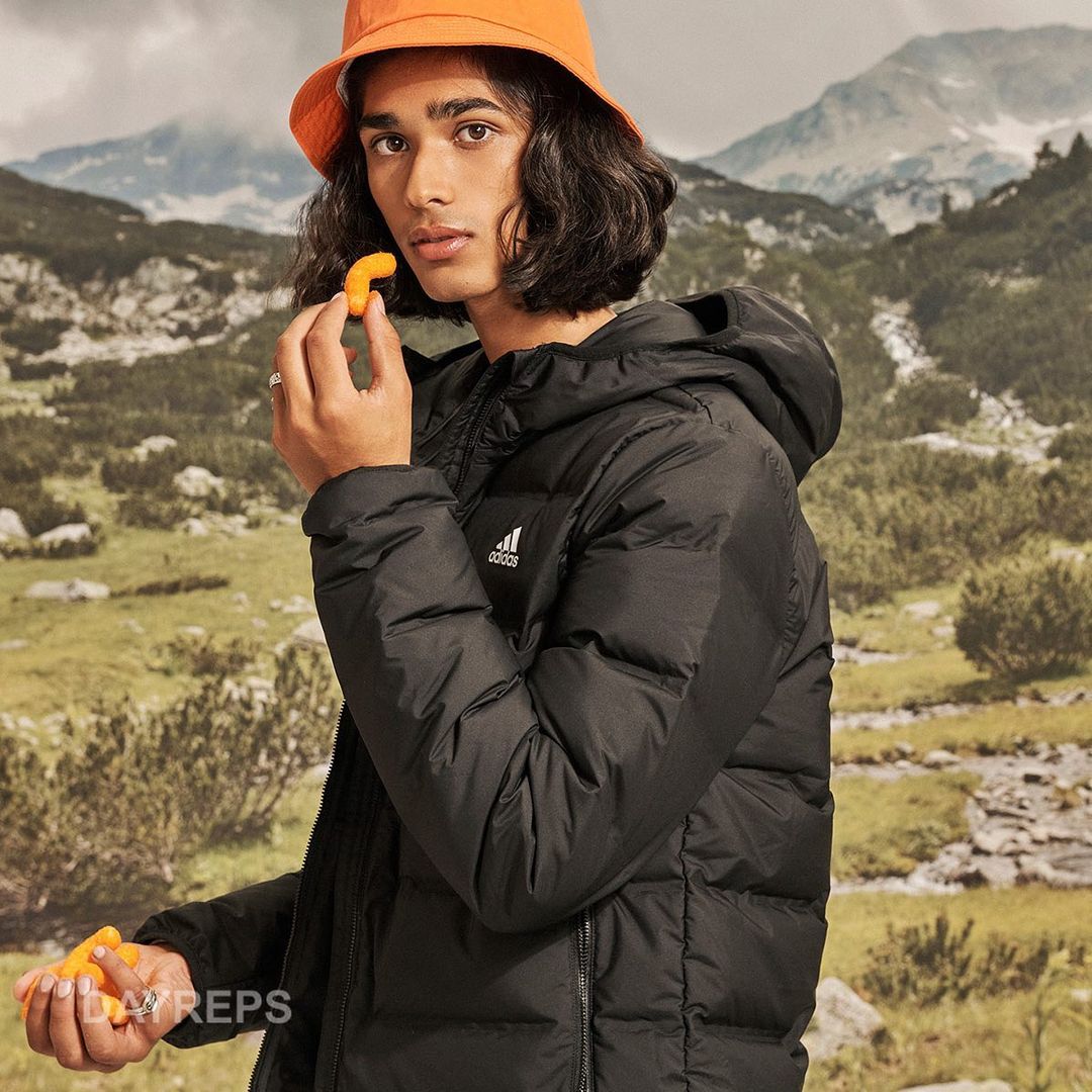 A young adult stands outdoors in a black puffer jacket and orange hat eating a snack with mountains and greenery in the background.