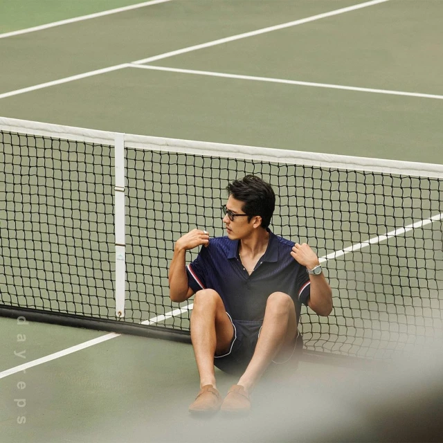 Man in polo shirt and shorts sitting on a tennis court near the net, wearing glasses and a wristwatch in natural light.