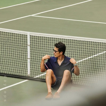 Man in polo shirt and shorts sitting on a tennis court near the net, wearing glasses and a wristwatch in natural light.