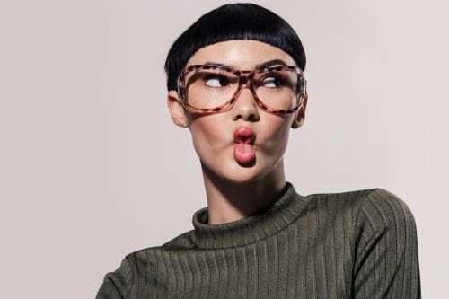 Attractive woman with short black hair and large glasses poses in front of a light background, sticking her tongue out.