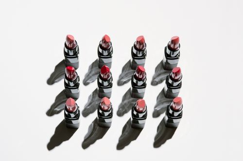 Overhead shot of a symmetrical grid of 12 lipsticks with varying shades of red and pink, casting strong, graphic shadows on white.