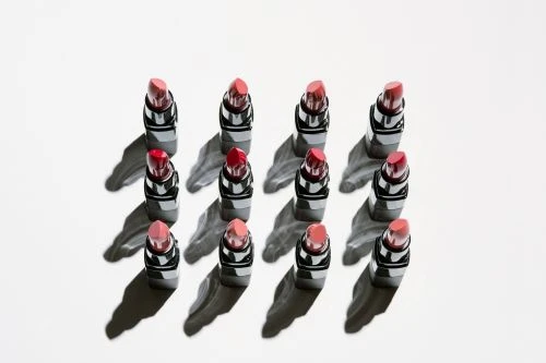 Overhead shot of a symmetrical grid of 12 lipsticks with varying shades of red and pink, casting strong, graphic shadows on white.