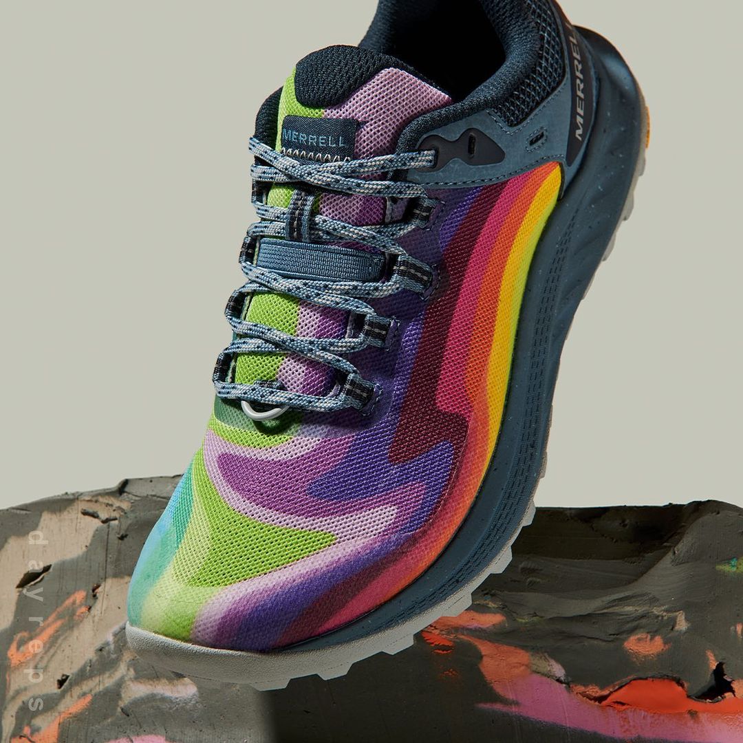 Merrell trail running shoe featuring a vibrant rainbow color pattern on a textured grey surface. Close-up product shot.