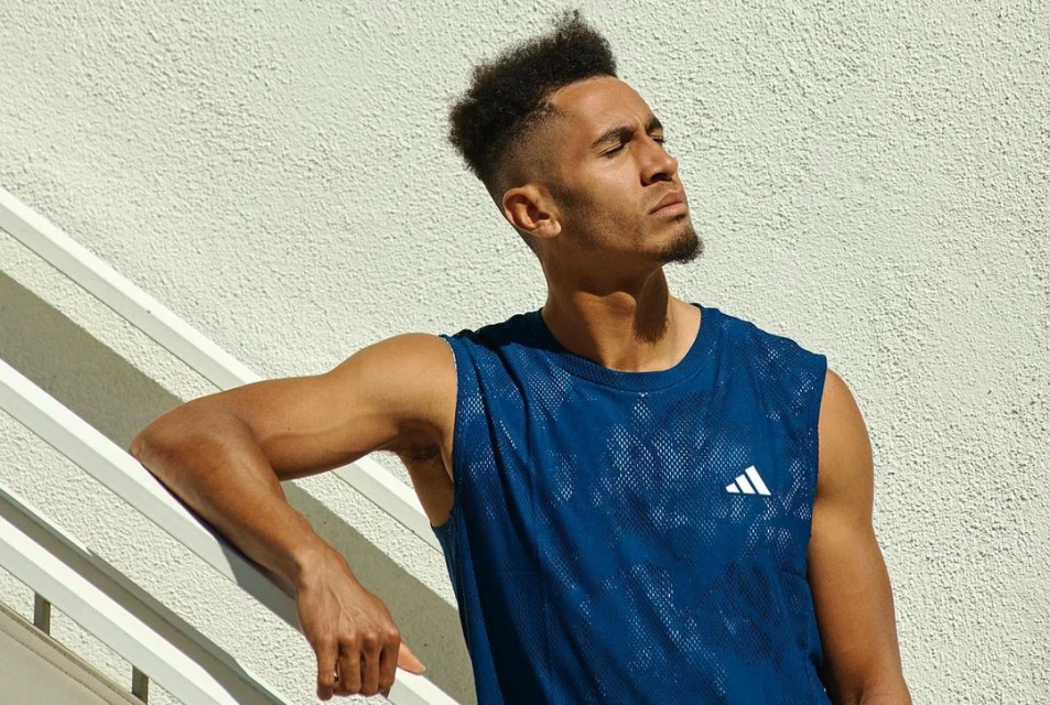 Athletic man with dark curly hair in blue mesh tank top leans against white railing outdoors in bright sunlight with closed eyes.