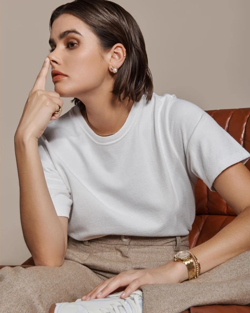 Portrait of a stylish woman with brown hair wearing a white t-shirt and herringbone pants, sitting on a leather chair in a studio.