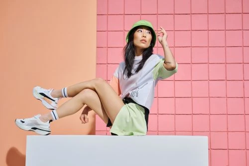 East Asian woman in green bucket hat and pastel sportswear sitting on a white block against a pink and peach tiled backdrop.