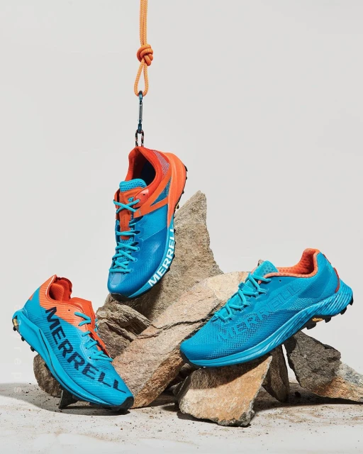 Three blue and orange Merrell running shoes are displayed on and hanging above a rock formation against a white backdrop in a studio setting.