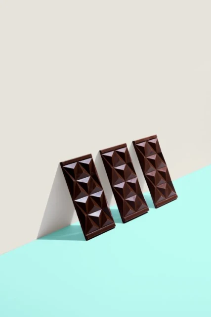Three chocolate bars with geometric pyramid pattern stand upright against a wall with two-tone off-white and aqua background.